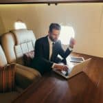 A business man in a suit works on his laptop at a table inside a private jet.