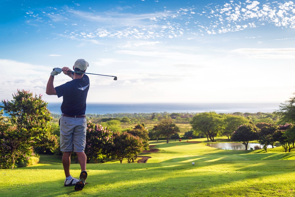 A man swings a golf club at a golf course near a large body of water